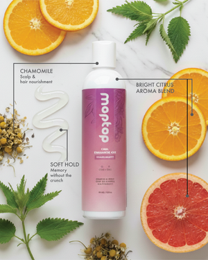 Bottle of Curl Enhancer Gel surrounded by Chamomile, Citrus slices, mint leaves, and clear cream